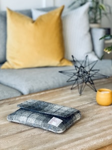 A cozy living room setting with a wooden coffee table. On the table, there is a grey patterned fabric pouch with a label, a small decorative object resembling a geometric star sculpture made of black metal, and a yellow candle holder. The background features a grey couch with a large mustard yellow pillow and a white pillow with grey stripes. A few green plants are visible, adding a touch of greenery to the scene.