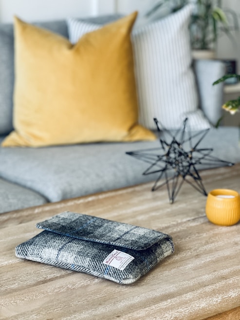 A cozy living room setting with a wooden coffee table. On the table, there is a grey patterned fabric pouch with a label, a small decorative object resembling a geometric star sculpture made of black metal, and a yellow candle holder. The background features a grey couch with a large mustard yellow pillow and a white pillow with grey stripes. A few green plants are visible, adding a touch of greenery to the scene.