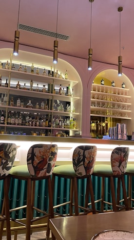 A stylish bar area featuring a well-stocked backlit shelf with a variety of liquor bottles. Tall, ornate bar stools with floral and bird patterns line the bar counter. The interior design includes warm lighting and a mix of pink and green tones, creating an elegant atmosphere.