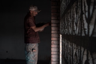 A man wearing a baseball cap is working with bricks, possibly constructing a wall, in a dimly lit environment. He appears focused on his task, holding a tool or object near the wall.