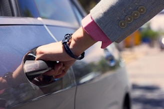 A hand holding a car door handle, wearing a business suit jacket with a pink shirt underneath and a black bracelet or watch.