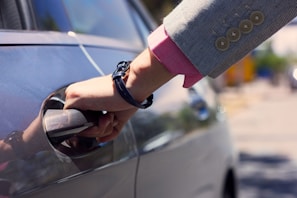 A hand holding a car door handle, wearing a business suit jacket with a pink shirt underneath and a black bracelet or watch.