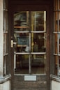 An old wooden door with glass panels that reveal a cluttered interior filled with shelves and various objects, suggesting a store or workshop. The door features a brass handle and a rectangular mail slot, with weathered wood and visible grain patterns.