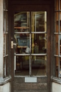 An old wooden door with glass panels that reveal a cluttered interior filled with shelves and various objects, suggesting a store or workshop. The door features a brass handle and a rectangular mail slot, with weathered wood and visible grain patterns.