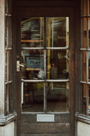An old wooden door with glass panels that reveal a cluttered interior filled with shelves and various objects, suggesting a store or workshop. The door features a brass handle and a rectangular mail slot, with weathered wood and visible grain patterns.