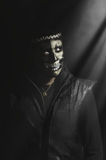 A person wearing dark clothing and a painted skull on their face stands under dim, moody lighting. A decorative headband adorns their head, and a chain necklace with a round pendant hangs around their neck. The atmosphere is shadowy and mysterious.