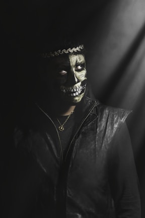 A person wearing dark clothing and a painted skull on their face stands under dim, moody lighting. A decorative headband adorns their head, and a chain necklace with a round pendant hangs around their neck. The atmosphere is shadowy and mysterious.