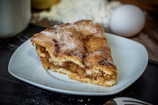A slice of apple pie is displayed on a white plate. The pie has a golden-brown crust sprinkled with sugar. The filling is visible, showing soft, caramelized apple pieces. In the background, there is an egg and some flour spilled on the surface, suggesting a baking setting.
