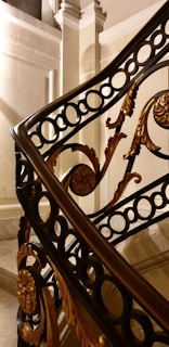 A close-up view of an ornate staircase railing featuring intricate designs. The railing is decorated with golden floral patterns and circular motifs within a black metal framework. The background shows a part of the staircase and walls with light beige paneling and molding details.