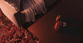 A cozy indoor setting with a beige sofa partially covered by a textured gray blanket. Next to the sofa is a wooden table holding two metal vases, casting soft shadows. The floor is covered in a thick, plush rug with a rich red and pink floral pattern, adding warmth and color to the scene.