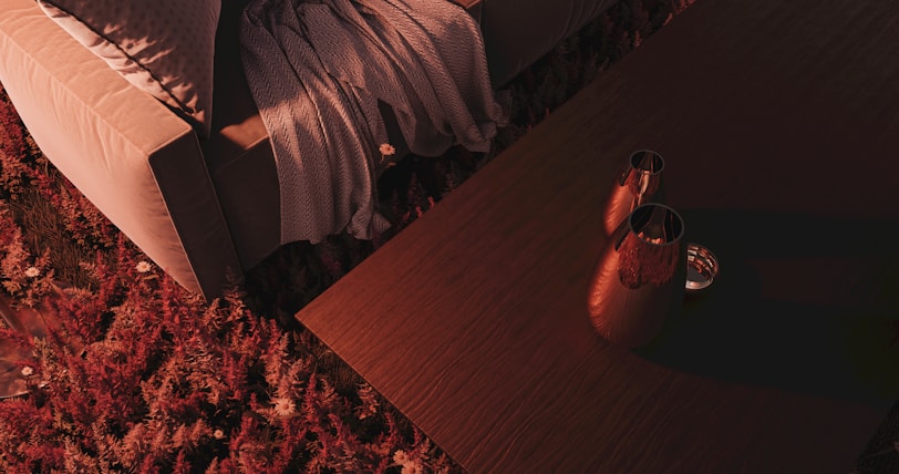 A cozy indoor setting with a beige sofa partially covered by a textured gray blanket. Next to the sofa is a wooden table holding two metal vases, casting soft shadows. The floor is covered in a thick, plush rug with a rich red and pink floral pattern, adding warmth and color to the scene.