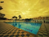 An outdoor swimming pool surrounded by a checkered tile deck. There are palm trees and buildings in the background under a cloudy, golden-hued sky. Pool ladders and floating lane dividers are visible in the pool.