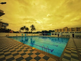 An outdoor swimming pool surrounded by a checkered tile deck. There are palm trees and buildings in the background under a cloudy, golden-hued sky. Pool ladders and floating lane dividers are visible in the pool.