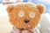A plush teddy bear with fuzzy light brown fur, featuring large button eyes and a round black nose. The bear's tongue is playfully sticking out, giving it a cute and whimsical expression. It wears a ribbon around its neck and is surrounded by a softly blurred background.