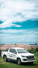 A white pickup truck with 'Test Driving' written on the side is parked on a grassy hillside. In the background, a line of peach and blue-roofed buildings is visible, along with a view of the ocean under a partly cloudy sky.