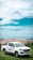 A white pickup truck with 'Test Driving' written on the side is parked on a grassy hillside. In the background, a line of peach and blue-roofed buildings is visible, along with a view of the ocean under a partly cloudy sky.