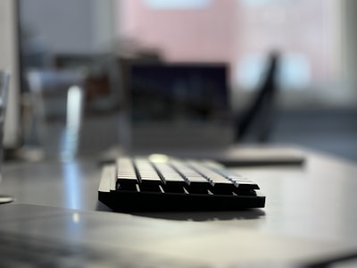 A close-up view of a keyboard on a desk, with a blurred background that appears to be an office environment. The keys on the keyboard are in focus, while the rest of the objects in the background, such as a chair and other desk items, are out of focus.