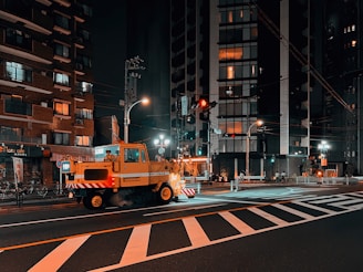 A nighttime urban street scene featuring a construction or utility vehicle parked on a crosswalk. Surrounding buildings are lit with various lights glowing from windows, suggesting residential or commercial spaces. Streetlights illuminate the area, with some reflected in the wet road surface, adding to the vibrant city atmosphere.