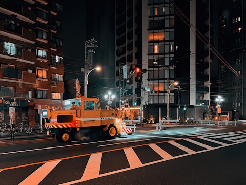 A nighttime urban street scene featuring a construction or utility vehicle parked on a crosswalk. Surrounding buildings are lit with various lights glowing from windows, suggesting residential or commercial spaces. Streetlights illuminate the area, with some reflected in the wet road surface, adding to the vibrant city atmosphere.