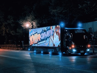 A truck parked on the side of the road at night, illuminated by streetlights. The side of the truck features a large, bright, colorful advertisement showcasing animated characters and Japanese text. The truck has glowing blue and orange lights, creating a vibrant contrast with the dark surroundings.