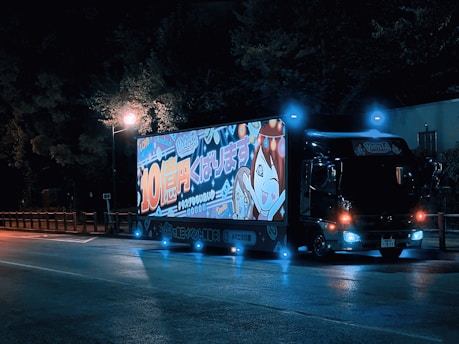 A truck parked on the side of the road at night, illuminated by streetlights. The side of the truck features a large, bright, colorful advertisement showcasing animated characters and Japanese text. The truck has glowing blue and orange lights, creating a vibrant contrast with the dark surroundings.