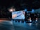 A truck parked on the side of the road at night, illuminated by streetlights. The side of the truck features a large, bright, colorful advertisement showcasing animated characters and Japanese text. The truck has glowing blue and orange lights, creating a vibrant contrast with the dark surroundings.