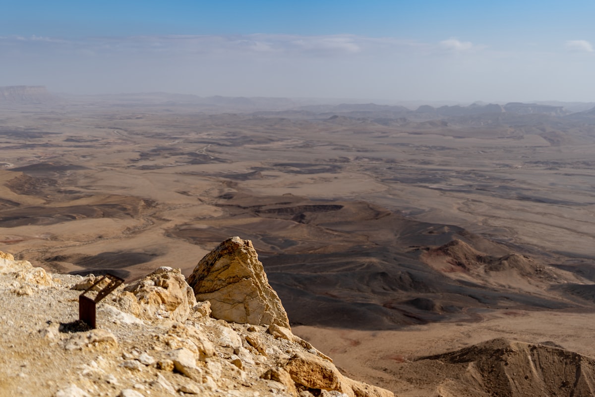 A view from a rocky cliff edge across an enormous desert basin stretching to the horizon in bright daylight