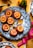 A decorative plate holds several baked desserts topped with caramelized orange slices and flower petals. Surrounding the plate are whole oranges with green leaves, star anise, and a metallic spoon. The tabletop has a mustard-yellow color with scattered spices and a pink ribbon.