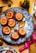 A decorative plate holds several baked desserts topped with caramelized orange slices and flower petals. Surrounding the plate are whole oranges with green leaves, star anise, and a metallic spoon. The tabletop has a mustard-yellow color with scattered spices and a pink ribbon.