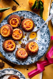 A decorative plate holds several baked desserts topped with caramelized orange slices and flower petals. Surrounding the plate are whole oranges with green leaves, star anise, and a metallic spoon. The tabletop has a mustard-yellow color with scattered spices and a pink ribbon.
