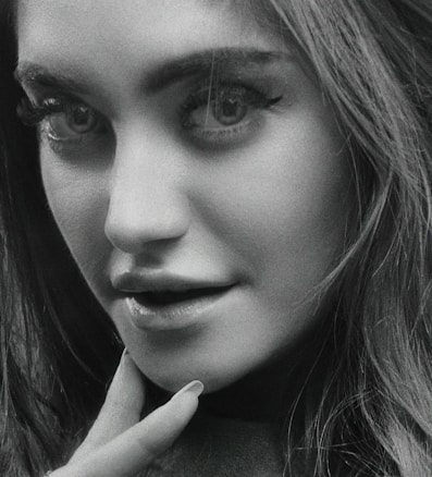 A close-up black and white portrait of a person with expressive eyes and soft lighting. The hair cascades down one side, and the focus is on the eyes and lips, with a hand gently touching the chin.
