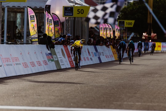 Cyclists are racing along a road lined with colorful banners and signs. A crowd of spectators is standing behind barriers, watching and cheering. The environment is festive with flags and markers indicating the race track.