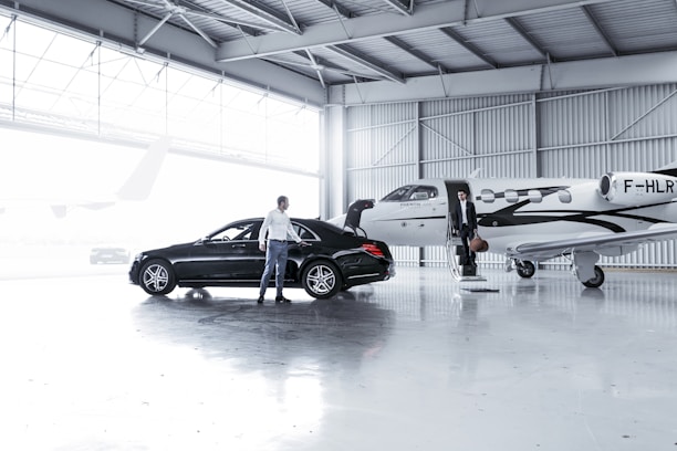 An indoor hangar with a sleek black car parked next to a small private jet. A person is standing next to the car, while another is descending the steps of the jet holding a bag. The hangar is well-lit and spacious with a large window offering natural light.