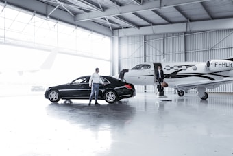 An indoor hangar with a sleek black car parked next to a small private jet. A person is standing next to the car, while another is descending the steps of the jet holding a bag. The hangar is well-lit and spacious with a large window offering natural light.