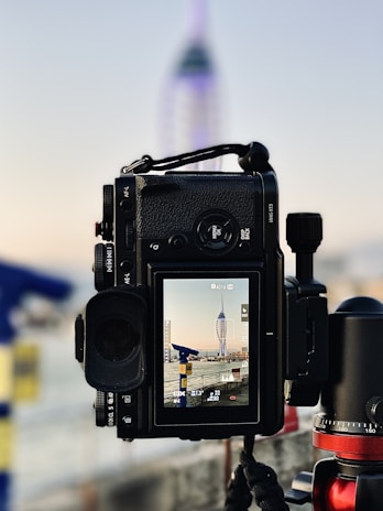 A camera on a tripod captures a tall, modern tower structure in the background. The camera's display shows a zoomed-in view of the tower, with details like exposure and focus settings visible. The background is blurred, with soft, pastel sky colors and parts of a waterfront or urban setting.