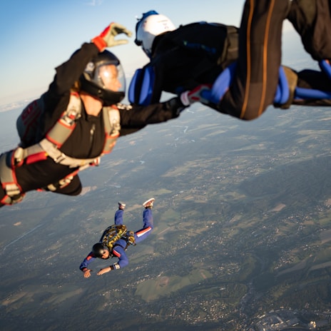 Three skydivers in mid-air, wearing helmets and jumpsuits equipped with parachute harnesses, float against a backdrop of sprawling landscapes far below. The lighting suggests it is late afternoon with clear skies.