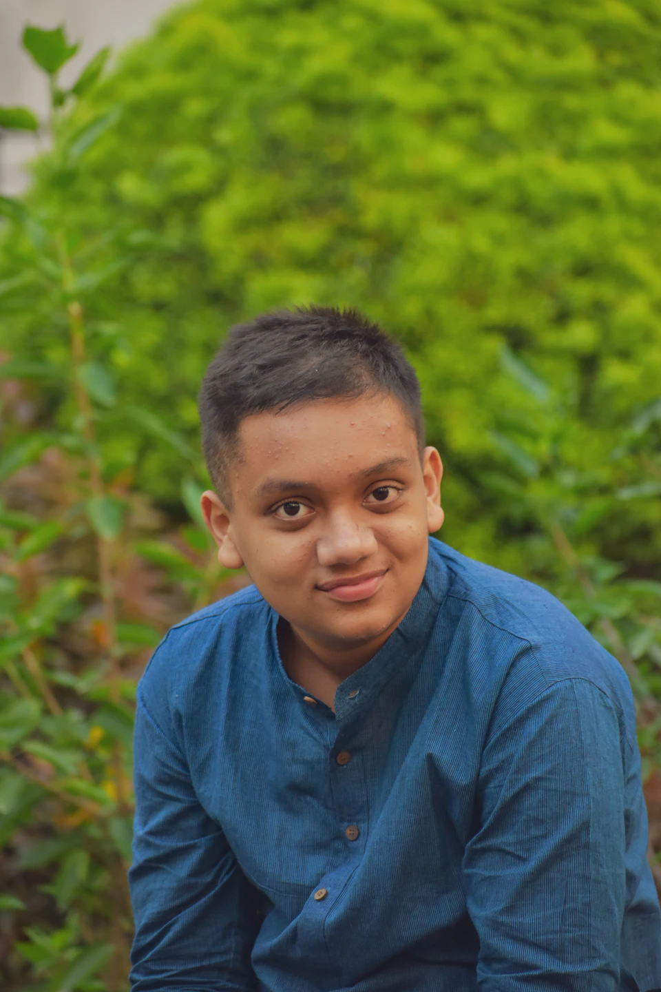 The image shows a young boy with short hair wearing a blue shirt, sitting on the ground and looking directly at the camera with a smile on his face. The background is blurred, suggesting an outdoor setting, possibly a park or garden. There are bushes in the background that add depth to the scene.