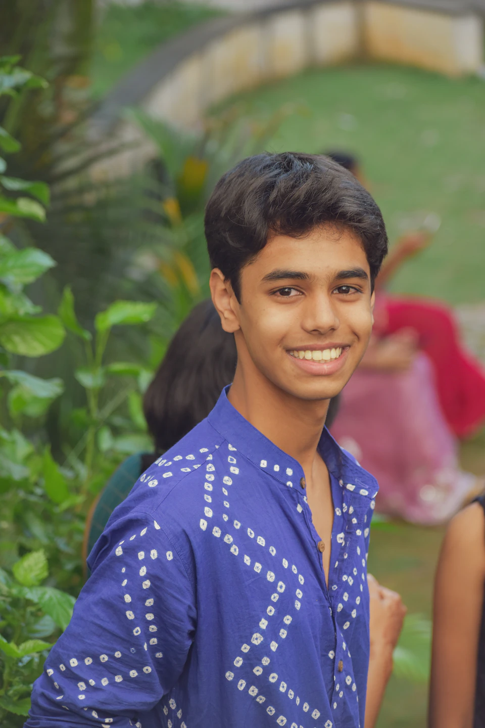 The image shows a young man with short dark hair wearing a blue shirt adorned with white dots, standing against a backdrop of lush green trees and bushes. The man is smiling warmly at the camera, creating an inviting atmosphere. In the background, there are two other people partially visible, adding depth to the scene.