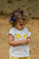A young child with curly hair and a blue bow, wearing a white shirt with a bright yellow butterfly design and patterned yellow shorts. The child appears focused on an object in their hands, set against a neutral, earthy background.
