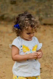 A young child with curly hair and a blue bow, wearing a white shirt with a bright yellow butterfly design and patterned yellow shorts. The child appears focused on an object in their hands, set against a neutral, earthy background.