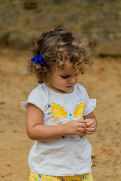 A young child with curly hair and a blue bow, wearing a white shirt with a bright yellow butterfly design and patterned yellow shorts. The child appears focused on an object in their hands, set against a neutral, earthy background.