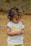 A young child with curly hair and a blue bow, wearing a white shirt with a bright yellow butterfly design and patterned yellow shorts. The child appears focused on an object in their hands, set against a neutral, earthy background.