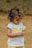 A young child with curly hair and a blue bow, wearing a white shirt with a bright yellow butterfly design and patterned yellow shorts. The child appears focused on an object in their hands, set against a neutral, earthy background.