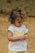 A young child with curly hair and a blue bow, wearing a white shirt with a bright yellow butterfly design and patterned yellow shorts. The child appears focused on an object in their hands, set against a neutral, earthy background.