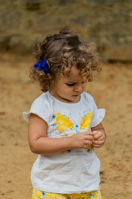 A young child with curly hair and a blue bow, wearing a white shirt with a bright yellow butterfly design and patterned yellow shorts. The child appears focused on an object in their hands, set against a neutral, earthy background.