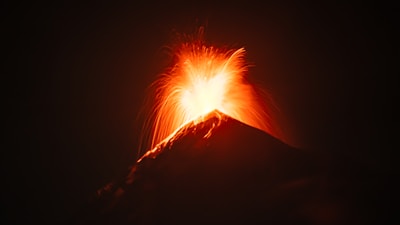 A dark landscape with a volcano actively erupting, spewing bright red and orange lava high into the night sky.