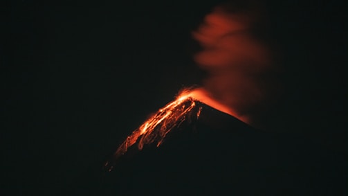 A volcano erupts against the night sky, casting a fiery glow. Streams of bright orange lava flow down the sides, and a thick plume of smoke billows upward.