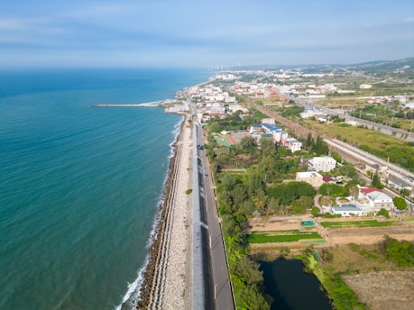 A coastal cityscape featuring a long road running parallel to the shoreline, bordered by a seawall made of rocks and concrete. The ocean appears calm with a rich blue hue. To the right of the road, a mix of residential buildings, green fields, and patches of vegetation stretches into the distance. The horizon is dotted with distant buildings and wind turbines, under a partly cloudy sky.