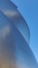 A striking and modern metallic building facade with curved, reflective surfaces. The structure's panels catch the light, producing subtle reflections and a sleek appearance. The clear blue sky serves as a backdrop, enhancing the building's futuristic aesthetic.