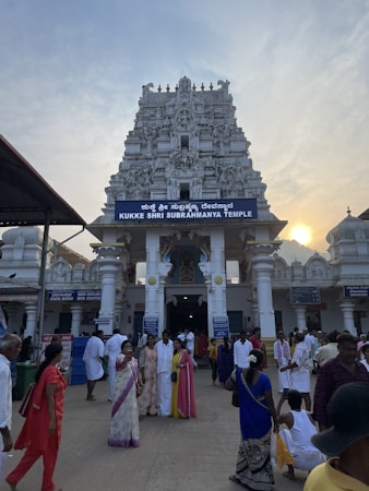 A temple with ornate architectural details is prominently featured, surrounded by a gathering of people, some dressed in traditional attire. The temple has a gopuram adorned with intricate sculptures. The sky is partly cloudy with the sun setting in the background, casting a warm glow.